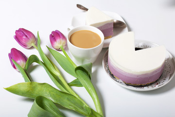 Blueberry cheesecake. A piece of ready-made dessert on a saucer. Jelly layers of different colors are visible. Near a cup of coffee and a bouquet of tulips.