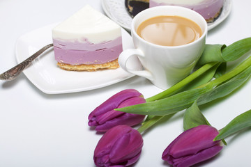 Blueberry cheesecake. A piece of ready-made dessert on a saucer. Jelly layers of different colors are visible. Near a cup of coffee and a bouquet of tulips.