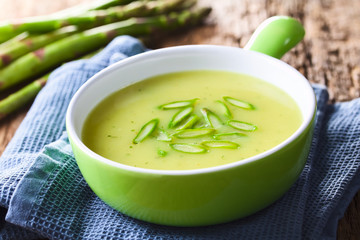 Cream of green asparagus soup in bowl, garnished with sliced asparagus on top, fresh asparagus in...