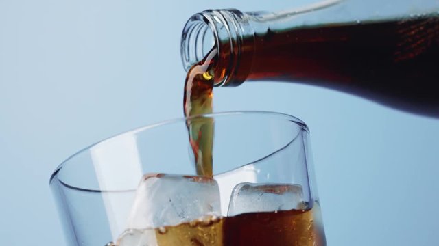 Closeup view from below of pouring cola into transparent glass with ice cubes on blue background