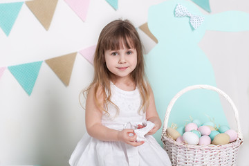 Easter 2019 Beautiful little girl in a white dress with Easter eggs and a basket. Big Easter eggs and bunnies, colorful place. A lot of different colorful Easter eggs. Multicolored flag, decor
