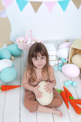 Easter! Little girl in overalls sits with a large Easter egg. Easter location, decorations. Family holidays, traditions. Colorful room. Child development.  Carrot, Easter decor, eggs