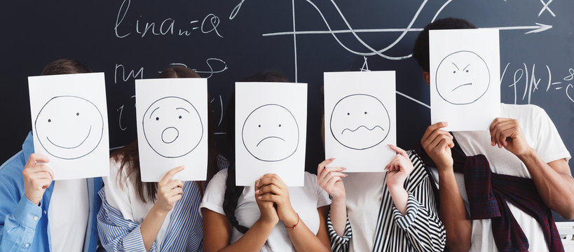 Friends holding papers with painted emotions in classroom - Powered by Adobe