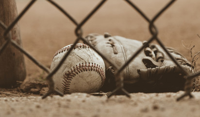 Old used baseball glove with ball on field behind dugout fence. - obrazy, fototapety, plakaty