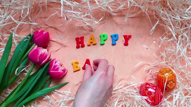 Woman put colourful words Happy Easter on table decorated with easter eggs, tulip flowers and decorative straw. Top view