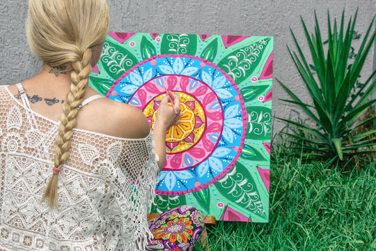 Painting outdoors, a young woman blonde draws a mandala on the nature sitting in the grass