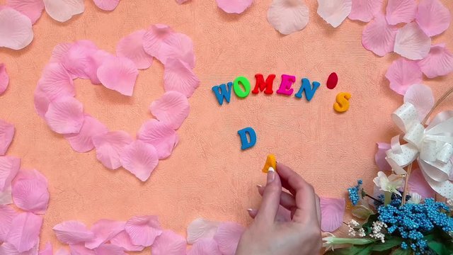 March 8 International Women's Day concept: woman put colourful letters of "Women's day" words next to heart made from pink rose petals and bouquet. 