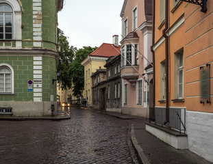 Fototapeta na wymiar Tallinn old city street in dull, murky rainy evening, with grey sky and wet cobblestone road. Old green, pink and orange buildings in the Old Tallinn, Estonia.