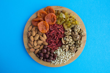 Dried apricots, raisins, goji berries ,different nuts on the round cutting board on the blue background.Top view.