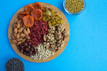 Dried apricots, raisins, goji berries,different nuts, chia seeds and  bee pollen on the round cutting board on the blue background.Top view.