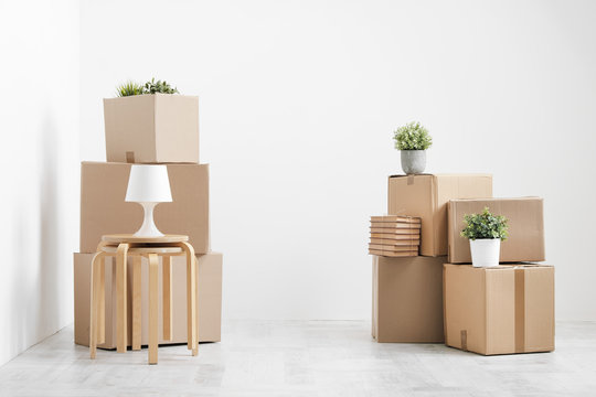 Cardboard boxes of things are stacked on the floor against a white wall. Books and table lamps and green plants in pots. The concept of moving to a new home.