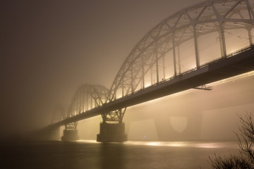 A mysterious evening fog above the river in big city. Bridge in the mist, cold weather scenery. Soft, blurry, misty look. Colorful, mystic industrial cityscape.