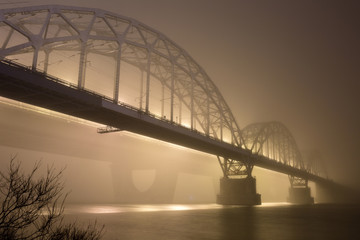 Obraz premium A mysterious evening fog above the river in big city. Bridge in the mist, cold weather scenery. Soft, blurry, misty look. Colorful, mystic industrial cityscape.