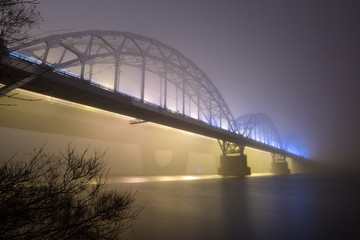 A mysterious evening fog above the river in big city. Bridge in the mist, cold weather scenery. Soft, blurry, misty look. Colorful, mystic industrial cityscape.