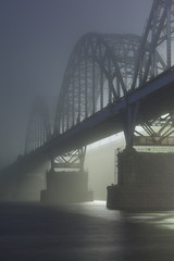 A mysterious evening fog above the river in big city. Bridge in the mist, cold weather scenery. Soft, blurry, misty look. Colorful, mystic industrial cityscape.