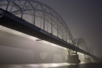 Obraz premium A mysterious evening fog above the river in big city. Bridge in the mist, cold weather scenery. Soft, blurry, misty look. Colorful, mystic industrial cityscape.
