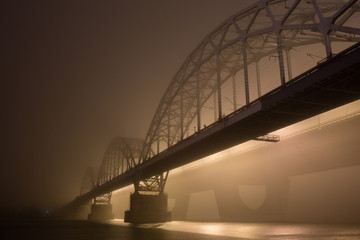 Obraz premium A mysterious evening fog above the river in big city. Bridge in the mist, cold weather scenery. Soft, blurry, misty look. Colorful, mystic industrial cityscape.