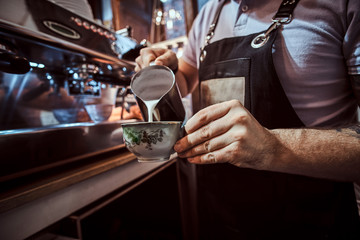 Barista in apron making a cappuccino, pouring milk in a cup in a restaurant or coffee shop