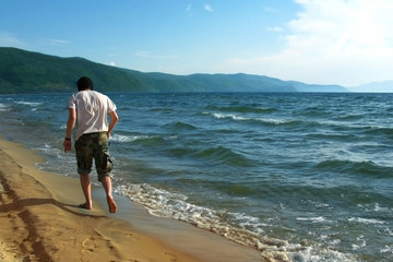 Alone young man walking on the beach barefoot. Sandy beach, mountains on the horizon, Sunny day.