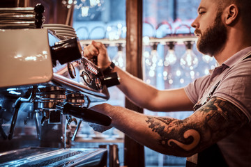 Handsome tattooed barista with stylish beard and hairstyle working on a coffee machine in a coffee shop or restaurant