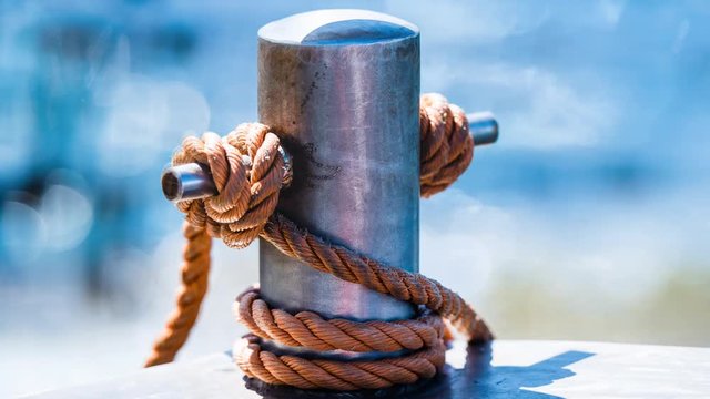 Abstract: Rope tied in a knot on a bollard. Mooring rope wrapped around the cleat on sea background. Metallic capstan in harbor for the mooring of yachts and boats with blurred background