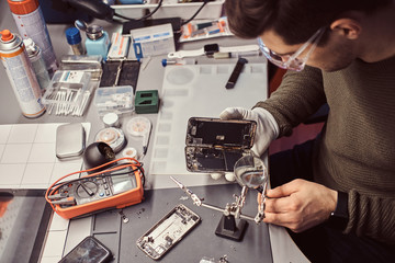 Serviceman uses magnifier and tweezers to repair damaged smartphone in the electronic workshop.