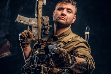 Special forces soldier wearing body armor holding assault rifle and aiming a gun at the camera. Studio photo against a dark textured wall