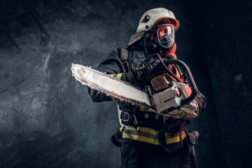 Firefighter in safety helmet and oxygen mask holding a chainsaw. Studio photo against a dark...