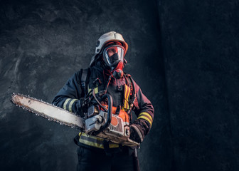 Obraz premium Firefighter in safety helmet and oxygen mask holding a chainsaw. Studio photo against a dark textured wall