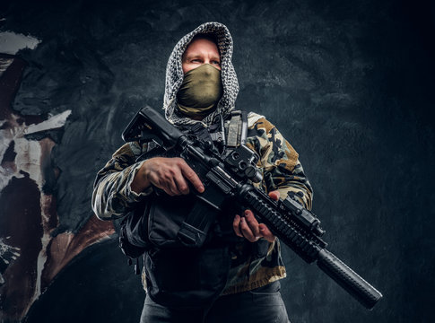 Special forces soldier in military uniform wearing mask and hood holding an assault rifle. Studio photo against a dark textured wall