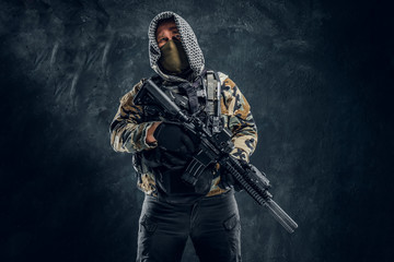 Fototapeta na wymiar Special forces soldier in military uniform wearing mask and hood holding an assault rifle. Studio photo against a dark textured wall