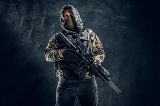 Special forces soldier in military uniform wearing mask and hood holding an assault rifle. Studio photo against a dark textured wall