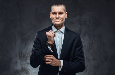 Elegantly dressed handsome man wearing a black classical suit, fastening buttons on the sleeves in dark studio.