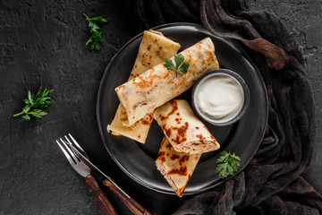 Crepes with chopped meat, greens and sour cream on black plate over dark background. Pancake week or Shrovetide. Healthy breakfast. Top view, flat lay