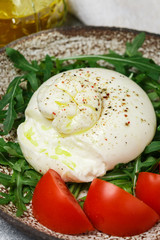 Sliced gourmet Italian cheese burrata served on a brown clay platter with fresh arugula, tomatoes, freshly ground pepper and olive oil with fork and knife. mozzarella and strachatella. Selective focus