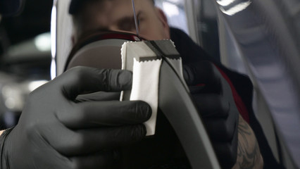 A professional worker guy (Male) applies ceramics (special liquid) to the car using an applicator (sponge) in black gloves and in a protective robe. Concept of: Auto service, Deteyling.