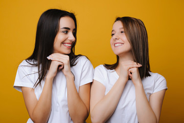 Portrait of two beautiful european female looking emotive and satisfied one to another laughing against a yellow background.