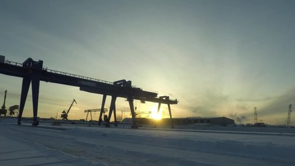 A large gantry crane standing still on the rail while automobile cranes worki behind it, time lapse. Gantry crane in the industrial zone in winter morning on rising sun background.