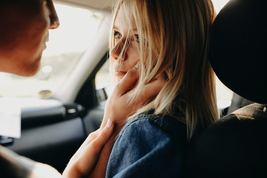 Amazing blonde woman sitting in the car with his boyfriend and looking at him while he is looking at her and holding with a hand her cheek while traveling.