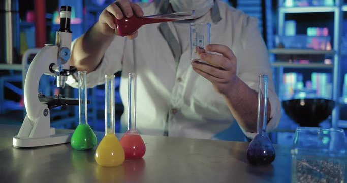 Scientist pours liquid from one flask to another. Microscopes and beakers with liquids stand on the table of a chemical laboratory.
