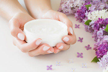 Young, perfect, groomed woman's hands holding moisturizing cream. Care about nails and clean, soft, smooth skin. Beautiful branches of fresh, colorful lilac flowers. Front view. Close up.