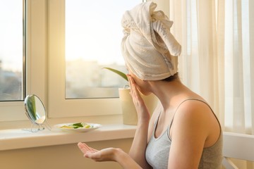Young woman at home near the window looking in mirror, caring for face with cream and making mask with cucumber and lemon, towel on head. Skin care, cosmetics, cosmetology, dermatology.