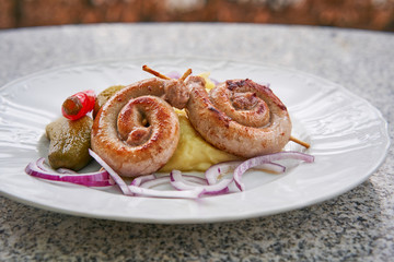 Obraz na płótnie Canvas Czech traditional and seasonal food. Spiral fried white wine sausages with mashed potatoes, pickles from cucumber and hot pepper, chopped fresh red onion served on garden table outdoor on white plate