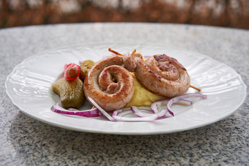 Czech traditional and seasonal food. Spiral fried white wine sausages with mashed potatoes, pickles from cucumber and hot pepper, chopped fresh red onion served on garden table outdoor on white plate
