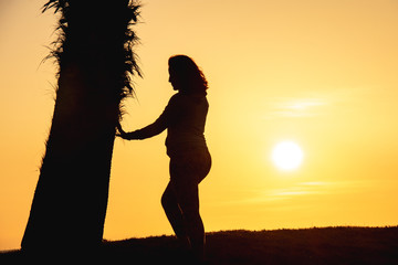 Fototapeta premium Pregnant woman touching her belly and watching sunset, next to a palm tree, at Almerimar, Almeria, Spain