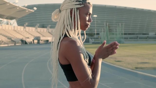 African sportswoman with braided blond hair walking across the running track rubbing her hands. Female feeling cold during training session in stadium. 