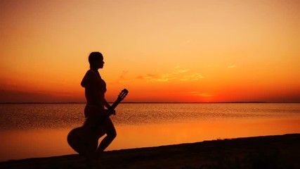Young girl walking with guitar on the beach at sunset