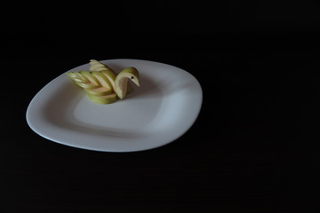 Table setting. The figure of a Swan from a green Apple on a white serving plate, stands on a table with a dark background.