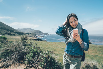 athletic asian woman traveler with backpack looking at smart phone while hiking in big sur mountain with blue sky. Nature along the route 101 at the California Coast from Los Angeles to San Francisco