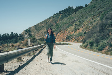 asian woman traveler with backpack walk on mountain road trip in 17 mile drive highway 1 california usa. full length of young girl hiker in sunglasses relax hiking in big sur seeing beautiful ocean